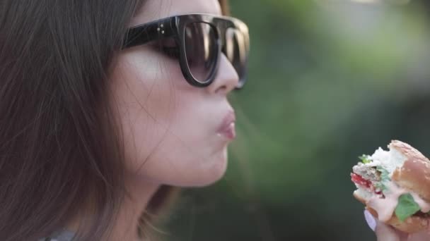 Девочка-подросток в стильных солнцезащитных очках с яркими губами наслаждается вкусным бургером
. - Кадры, видео