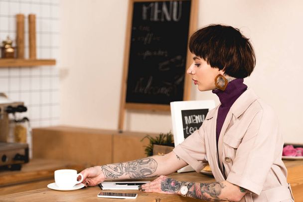 attractive businesswoman sitting at bar counter near coffee cup, notebook and smartphone - Foto, immagini