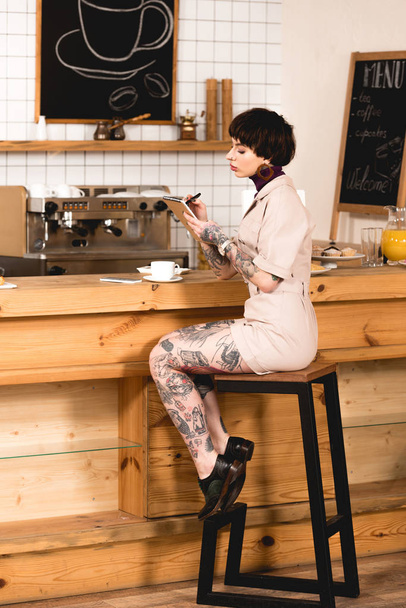 fashionable, tattooed businesswoman sitting at bar counter and writing in notebook - Photo, Image