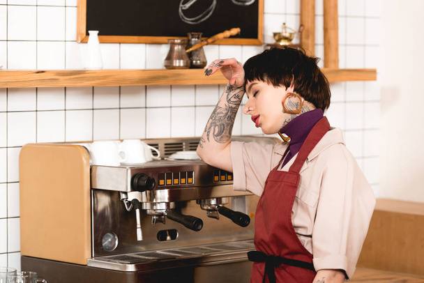 pretty, exhausted barista standing near espresso machine in cafeteria - Photo, Image