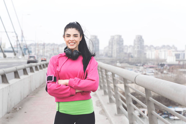 Привлекательная красивая брюнетка с дружелюбной улыбкой в спортивной одежде, стоящая на мосту города и смотрящая в камеру
 - Фото, изображение