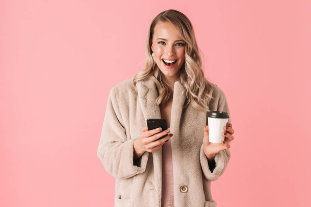 Happy young pretty woman posing isolated over pink wall background using mobile phone drinking coffee. - Photo, image