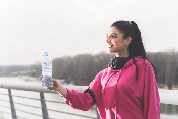attraktive, fröhliche, sportliche junge Frau steht auf einer Brücke am Fluss und nimmt eine Plastikflasche Wasser - Foto, Bild