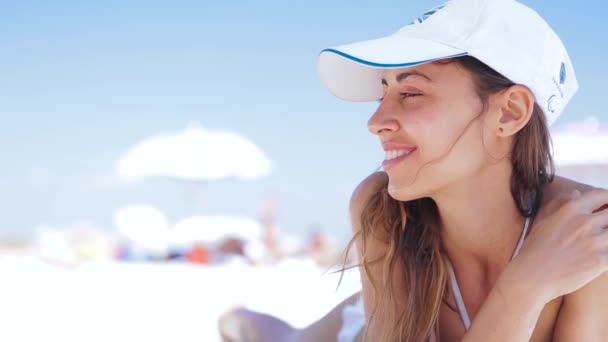 beautiful tanned smiling woman is lying on the beach on a white sand, relaxing and listen music from the phone with small headphones. slim sexy woman in bikini and white cap enjoing and suntanning on - Footage, Video
