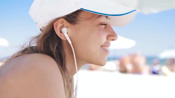 zaostřená portrét nádherné, vyčiněné usměvavé ženy leží na pláži na bílém písku, odpočívá a naslouchá hudbě z telefonu s malými sluchátky. štíhlá sexy žena v bikinách a bílé čepici - Záběry, video
