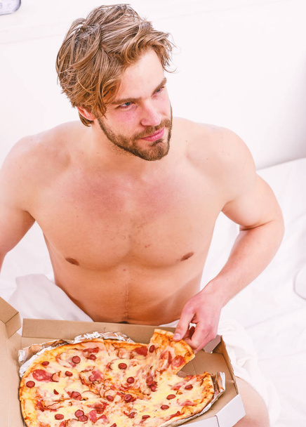 Portrait of lazy muscular man eating pizza while laying on a bed at home. Cropped image of shirtless sexy man with pizza on bed. Break diet concept. - Photo, Image