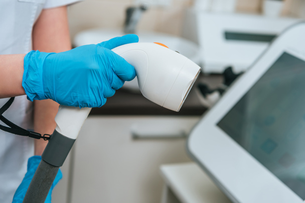 Weergave van de schoonheidsspecialist in rubberen handschoenen houden laser apparaat in kliniek bijgesneden - Foto, afbeelding