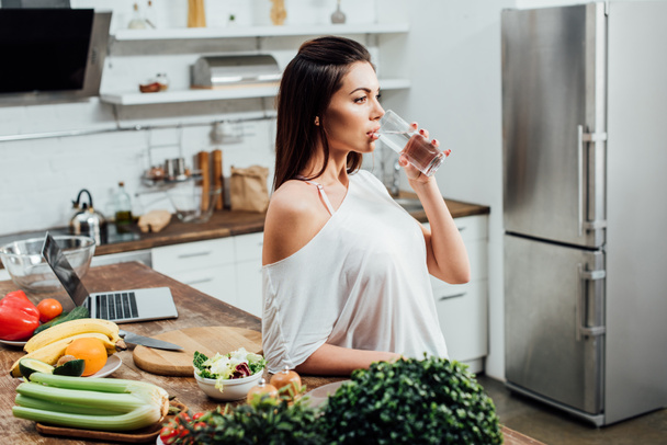 nachdenkliche attraktive Frau trinkt Wasser am Tisch in der Küche - Foto, Bild