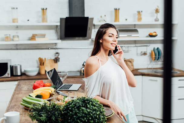 Happy young woman talking on smartphone with smile in kitchen - Photo, Image
