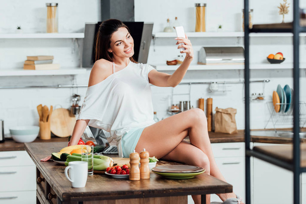 Glimlachend meisje zittend op houten tafel en het nemen van selfie in de keuken - Foto, afbeelding