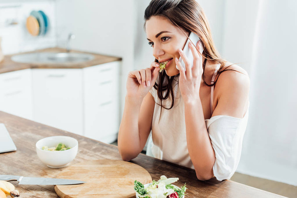 Hezká dívka pojídáním salátu a mluvení na smartphone v kuchyni - Fotografie, Obrázek