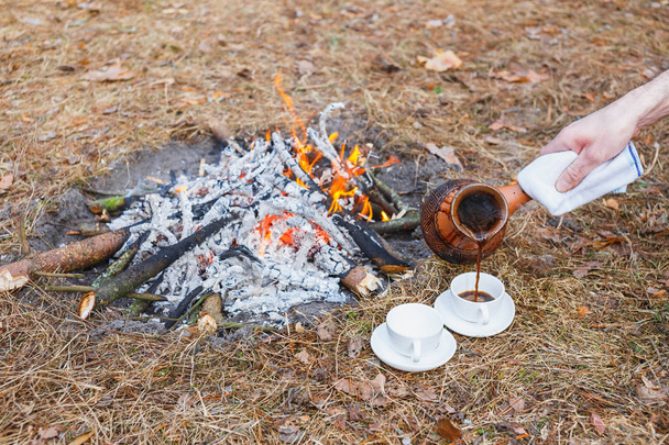 A man pours coffee from an earthen Turkish pot into small white cups on a campfire in the spring forest. - 写真・画像