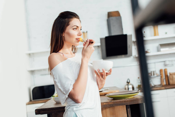 Dreamy woman holding bowl and eating cut fruit in kitchen - Photo, Image