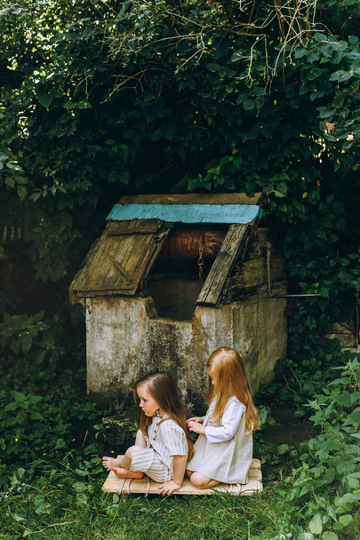 two beautiful girls in white antique shirts near a well on a background of grass and trees - Foto, imagen