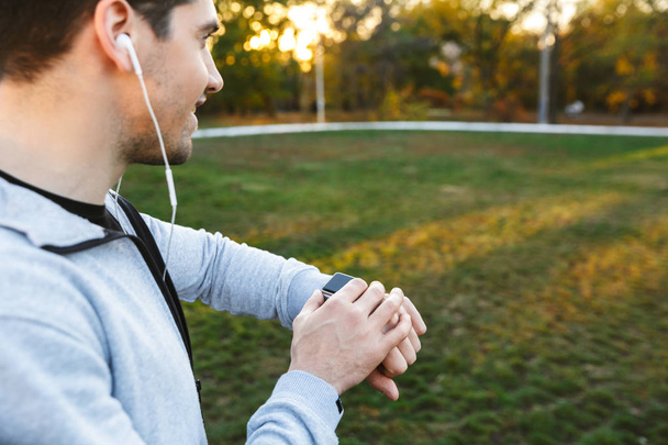 Handsome young sportsman outdoors in park listening music with earphones looking at watch. - Photo, Image