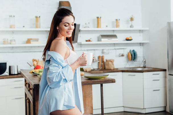 Smiling sexy girl in white lingerie holding cup of coffee in kitchen - Photo, Image