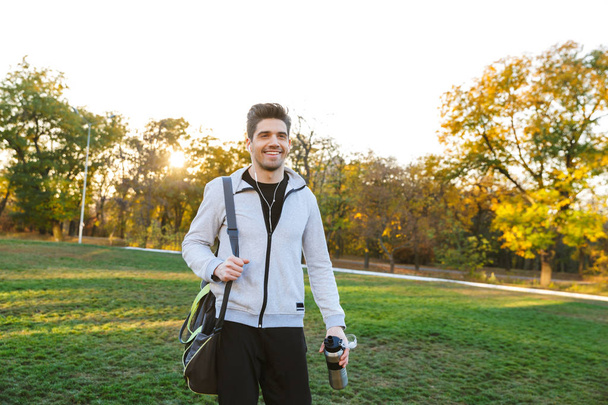 Sportsman outdoors in park listening music with earphones walking with bag. - Foto, immagini