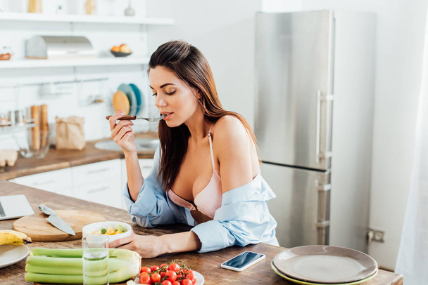 Сексуальная девушка в лифчике и рубашке ест фруктовый салат на кухне
 - Фото, изображение
