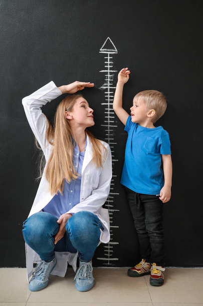 Γυναίκα γιατρός με μικρό αγόρι μέτρηση ύψους κοντά στο σκοτάδι τείχος - Φωτογραφία, εικόνα