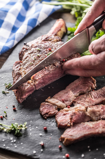 Freshly grilled tomahawk steak on slate plate with salt pepper rosemary and parsley herbs. Chef with knife cuts juicy bovine steak - 写真・画像