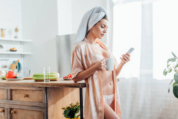 Sexy girl in lingerie and housecoat with towel on head holding cup of coffee and using smartphone in kitchen - Foto, Imagem
