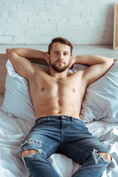 good-looking and muscular man in jeans lying on bed and looking at camera in bedroom  - Фото, зображення
