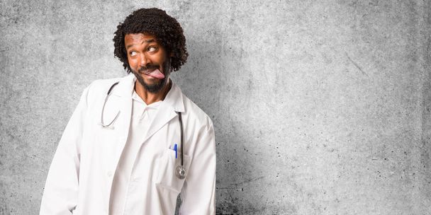 Bonito médico afro-americano expressão de confiança e emoção, divertido e amigável, mostrando a língua como um sinal de jogo ou diversão
 - Foto, Imagem