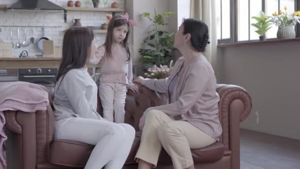 Grandmother, mother and little daughter spend time together sitting on the sofa in modern apartment. The girl standing on the cout, mom and granny looking at her smiling. Concept of generation. - Séquence, vidéo