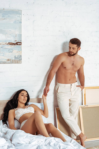 muscular and good-looking man in trousers holding hand with beautiful woman in lace bra  - Φωτογραφία, εικόνα