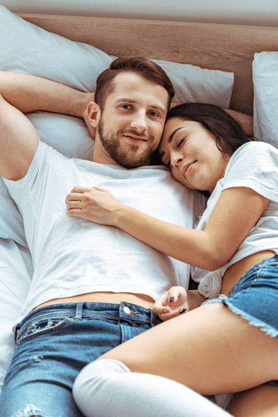 knappe man in spijkerbroek en mooi en lachende vrouw knuffelen in slaapkamer  - Foto, afbeelding