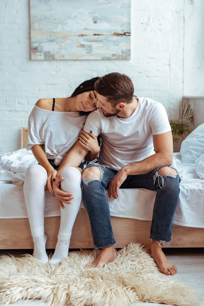 pohledný muž v džínách a krásná žena sedí na posteli a objímání  - Fotografie, Obrázek