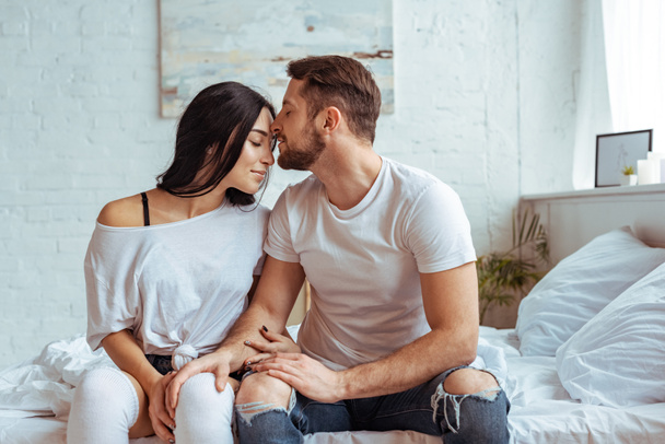 handsome man in jeans and beautiful woman sitting on bed and hugging  - Zdjęcie, obraz
