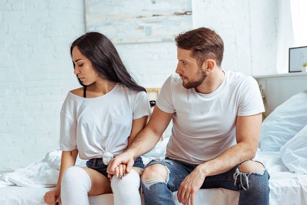 handsome man in jeans holding hand of beautiful and sad woman in t-shirt sitting on bed  - Foto, afbeelding