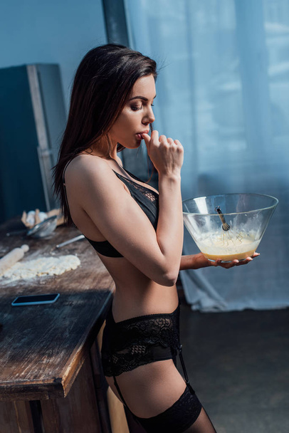 Vista lateral de la joven sexy en lencería negra lamiendo el dedo mientras batiendo huevos con batidor en la cocina
 - Foto, Imagen