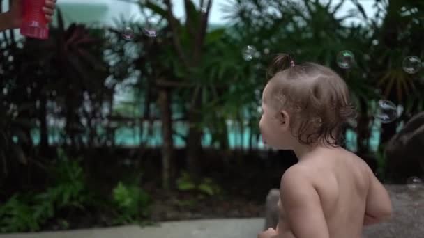 Mutter mit Tochter pustet Seifenblasen im Garten am Pool, Tochter freut sich und hebt die Hände zu den Seifenblasen, Zeitlupe - Filmmaterial, Video