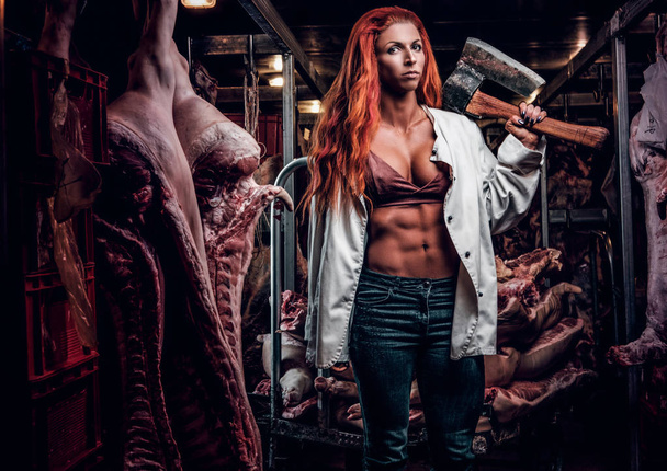 Mulher fitness ruiva vestindo camisa branca desabotoada posando com machado em um armazém refrigerado no meio de carcaças de carne
 - Foto, Imagem