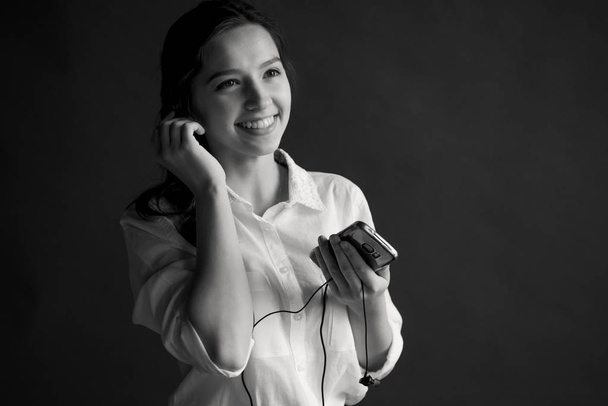 young pretty girl with brilliant hair with a nice smile, dressed in a white shirt on a gray background, holds small headphones for music and the phone and listens to the music - Zdjęcie, obraz