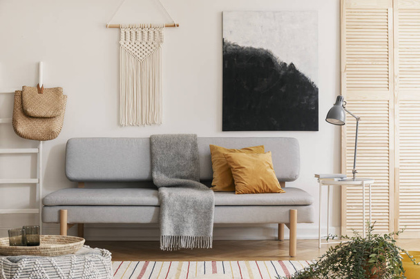 Abstract black and white painting and handmade macrame on white wall of natural living room interior with grey fashionable couch with yellow pillow and warm blanket - Fotografie, Obrázek