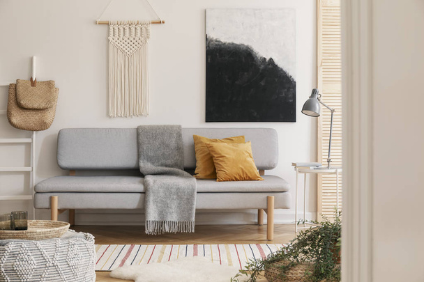 White handmade macrame above grey comfortable sofa with orange pillows and warm blanket, real photo with mockup on the empty wall - Foto, afbeelding