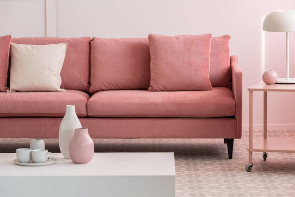 Pastellrosa und weiße Vasen und Kaffeebecher auf kleinem Tisch im rosafarbenen Wohnzimmer mit bequemem Sofa - Foto, Bild
