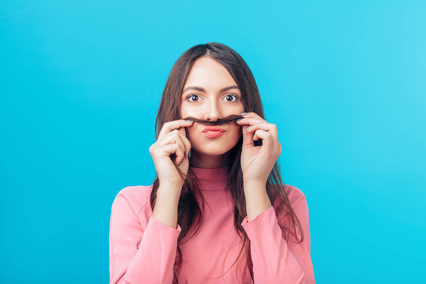 Portrait of young woman having fun shows moustache hair on blue background. Humor concept  - Photo, image