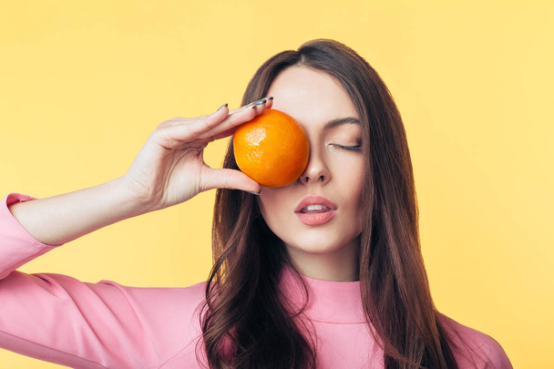 Beautiful young woman holding orange in front of eye on yellow background. Healthy food concept   - Photo, image