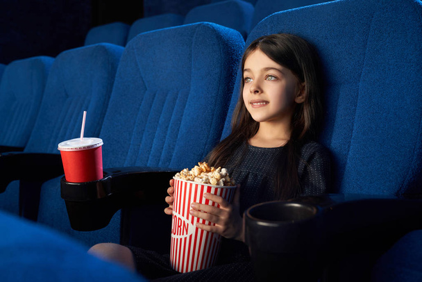 Cute kid sitting with popcorn bucket in cinema. - Zdjęcie, obraz
