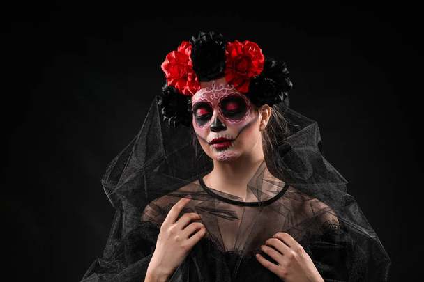 Young woman with painted skull on her face for Mexico's Day of the Dead against dark background - Photo, Image