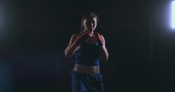 Prachtige vrouwelijke bokser otbryvatyvat klappen voert een strijd met schaduw in een donkere hall van de hall in slow motion in blauwe kleding en rode pleisters op haar polsen. vliegen rond de - Video