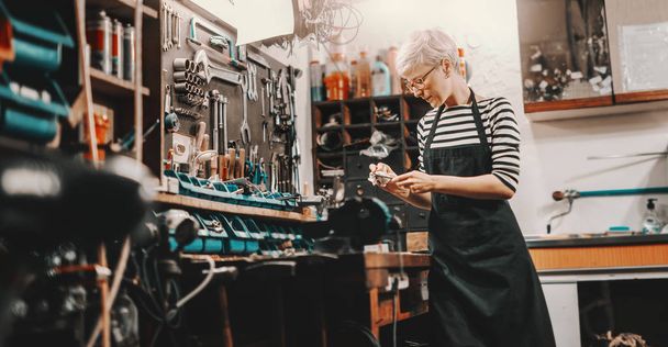 Beautiful Caucasian female worker with short blonde hair and eyeglasses taking tool fromm wall to repair bicycle. Bike workshop interior. - Photo, Image