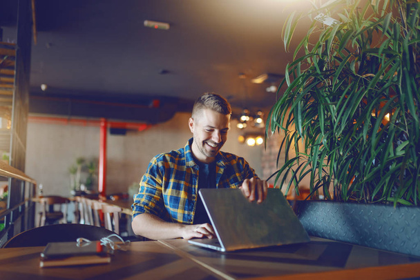 Przystojny uśmiechający się freelancer zarośnięty Kaukaska w plaid shirt otwarcie laptopa siedząc w kawiarni. - Zdjęcie, obraz