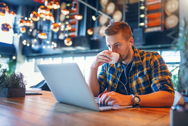 Giovane blogger caucasico con auricolari nelle orecchie e camicia a quadri che beve caffè e usa il computer portatile mentre è seduto in caffetteria
. - Foto, immagini