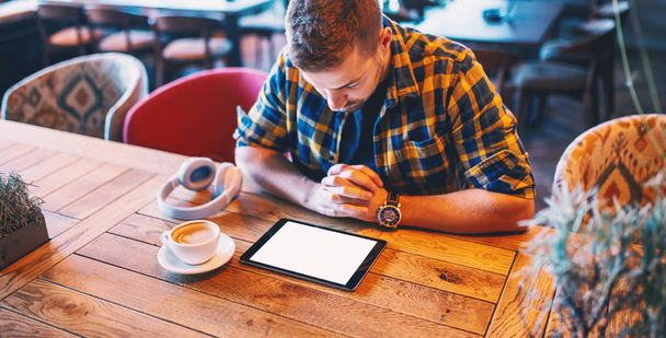 Handsome Caucasian man in plaid shirt holding hands on table and looking at tablet. On table headphones and coffee, coffee shop interior. - Valokuva, kuva