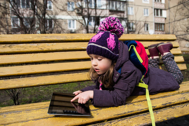 Small girl is lying on a bench outdoors and touches digital tabl - Photo, Image